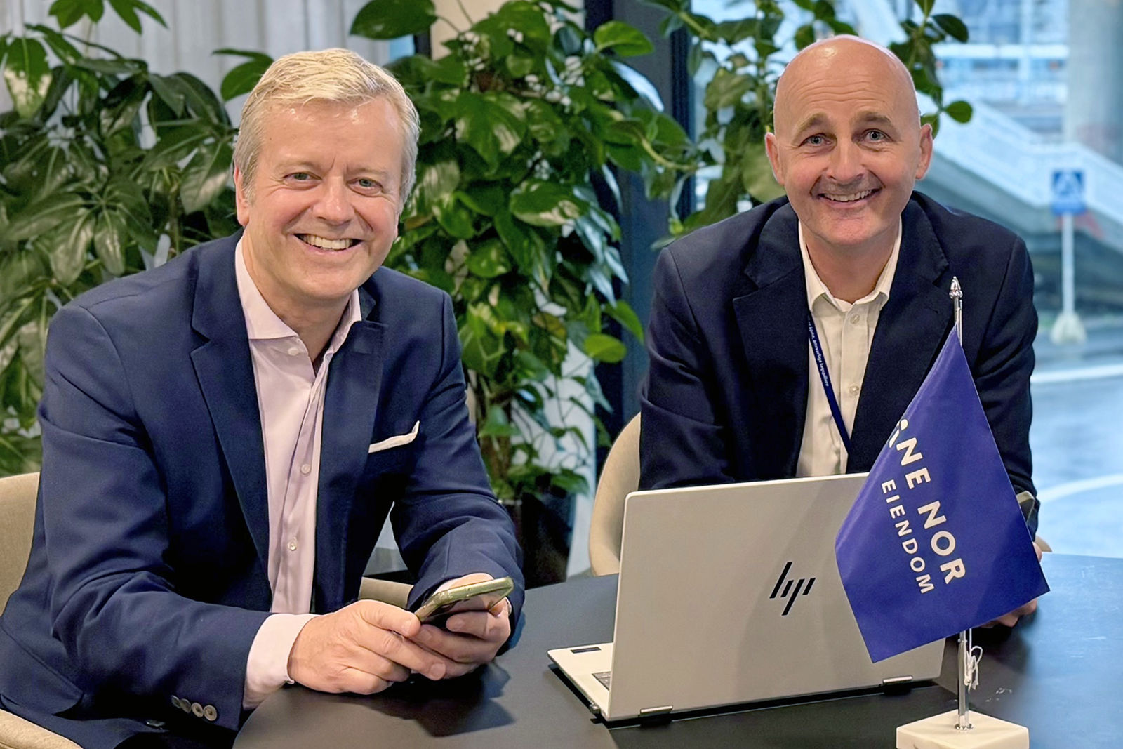 Personene sitter smilende ved et bord med en PC og et Bane NOR-flagg.