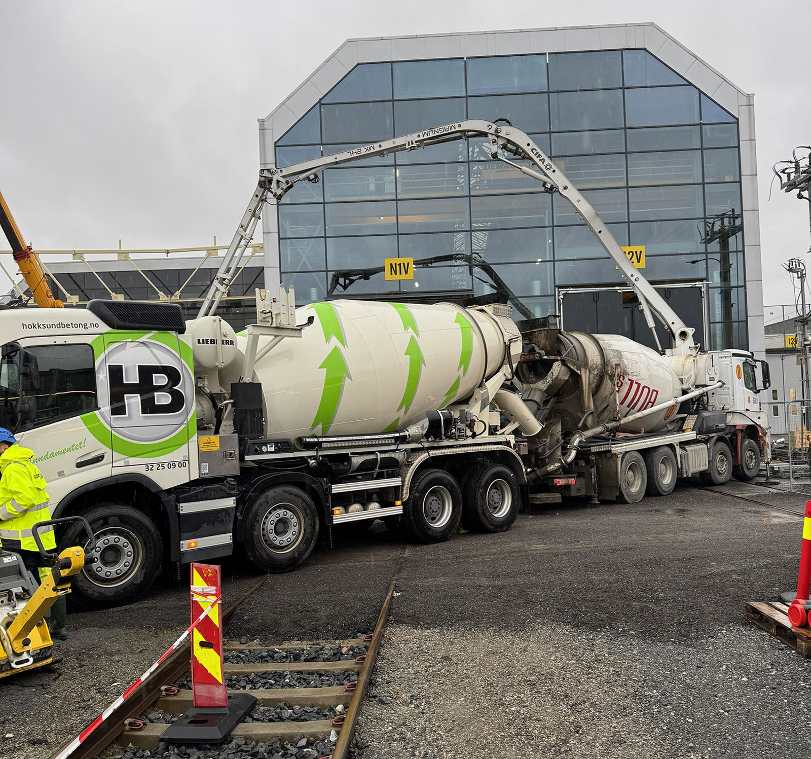 To betongbiler med betongpumper foran eksisterende toghall. Et jernbanespor i forgrunnen.